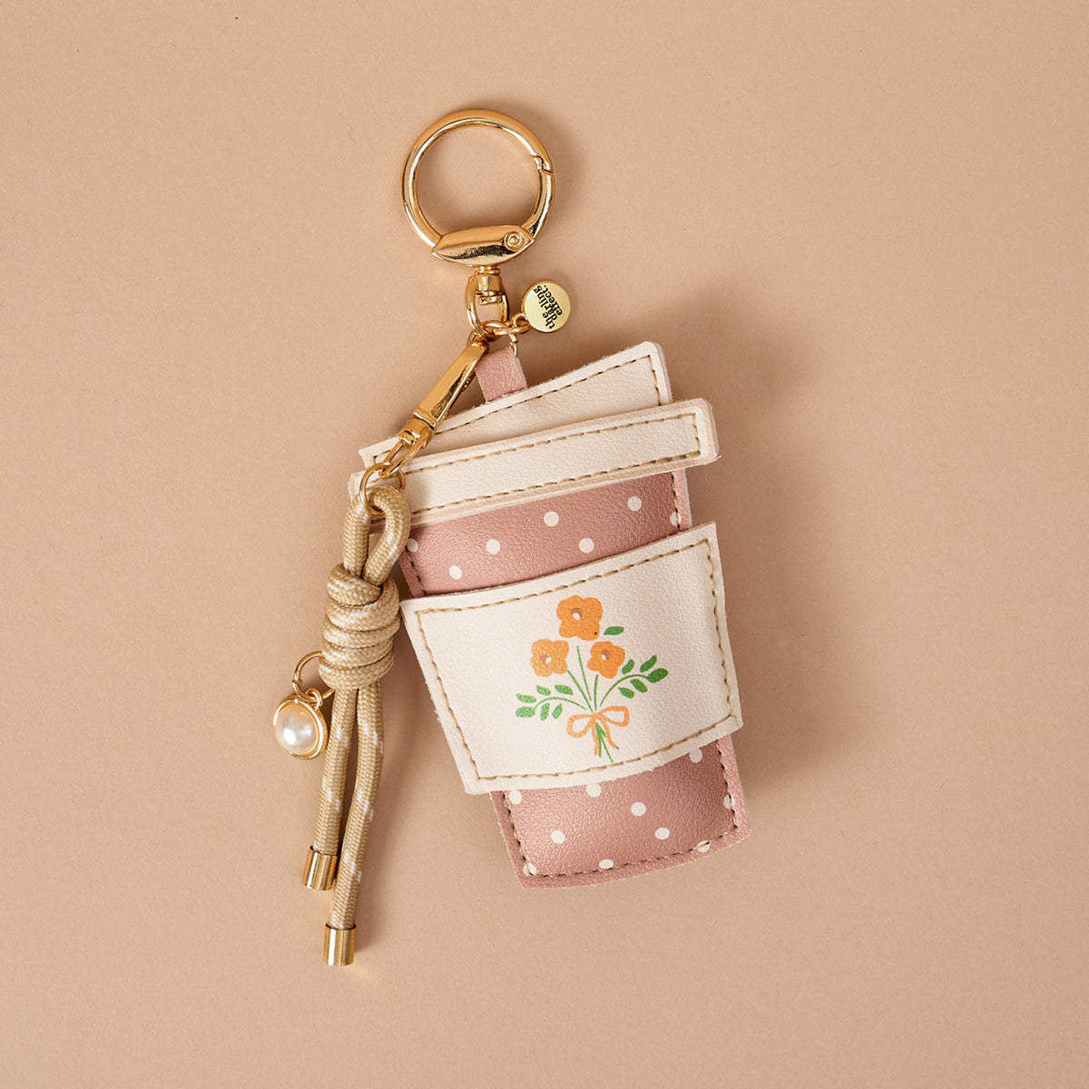 The Darling Effect’s Coffee Run Vegan Leather Bag Charm—shaped like a pink polka-dotted coffee cup with a white sleeve adorned with orange flowers, a gold key ring, and a knotted cord with a pearl charm—makes the perfect playful accessory for your bag. Beautifully displayed on a beige background, this charming piece is an ideal choice for anyone seeking fun Vegan Leather Bag Charms.