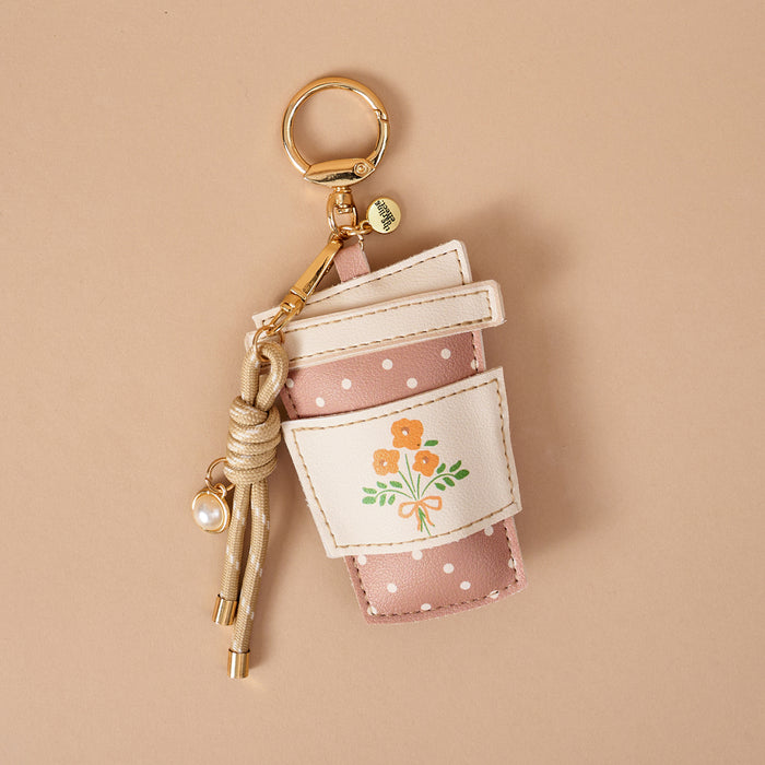 The Darling Effect’s Coffee Run Vegan Leather Bag Charm—shaped like a pink polka-dotted coffee cup with a white sleeve adorned with orange flowers, a gold key ring, and a knotted cord with a pearl charm—makes the perfect playful accessory for your bag. Beautifully displayed on a beige background, this charming piece is an ideal choice for anyone seeking fun Vegan Leather Bag Charms.