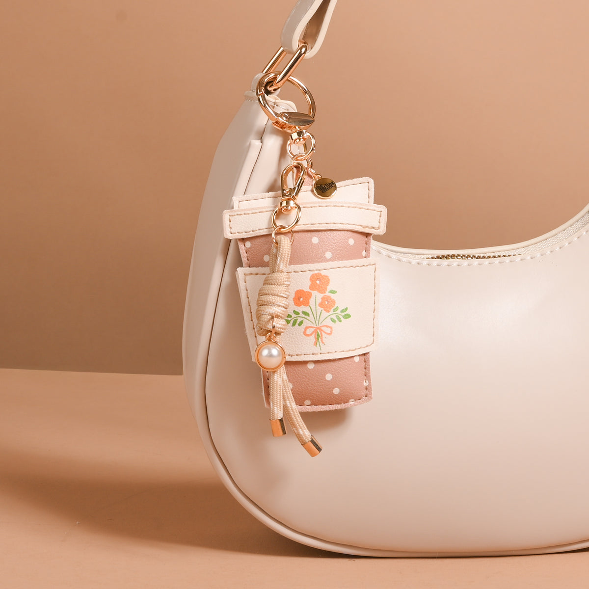 A cream-colored handbag is adorned with The Darling Effect’s Coffee Run Vegan Leather Bag Charm—a playful pink floral keychain and a small pouch featuring an orange flower and polka dots, both hanging from a gold clasp on the handle. These charming vegan leather accents elevate the stylish accessory, showcased against a beige background.
