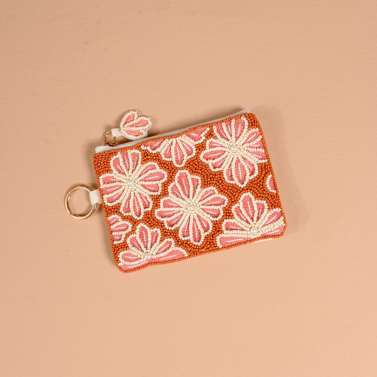 The Sienna Spice Handcrafted Beaded Pouch by The Darling Effect features a floral pattern in shades of pink and white on an orange background, complete with a zipper and attached keyring, and is displayed on a light beige surface.