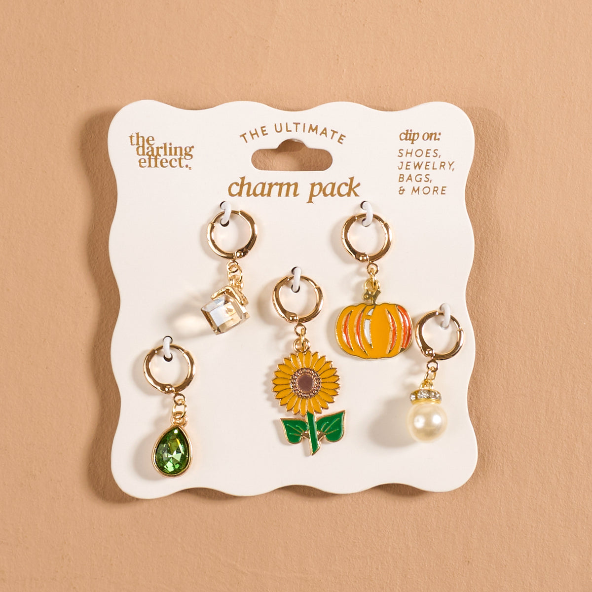 Introducing the Sunflower Fields The Ultimate Charm Pack by The Darling Effect—a stylish set of six gold clip-on accessories featuring a clear gem, orange pumpkin, green gem, sunflower, pearl, and golden droplet. Perfect for adding a touch of elegance to your jewelry collection. Beautifully displayed on a white card against a beige background.