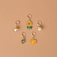 Sunflower Fields The Ultimate Charm Pack by The Darling Effect features five gold keychains—a white house, a sunflower, a pearl, a green teardrop gem, and an orange pumpkin—beautifully arranged on a tan background in a loose circular pattern.