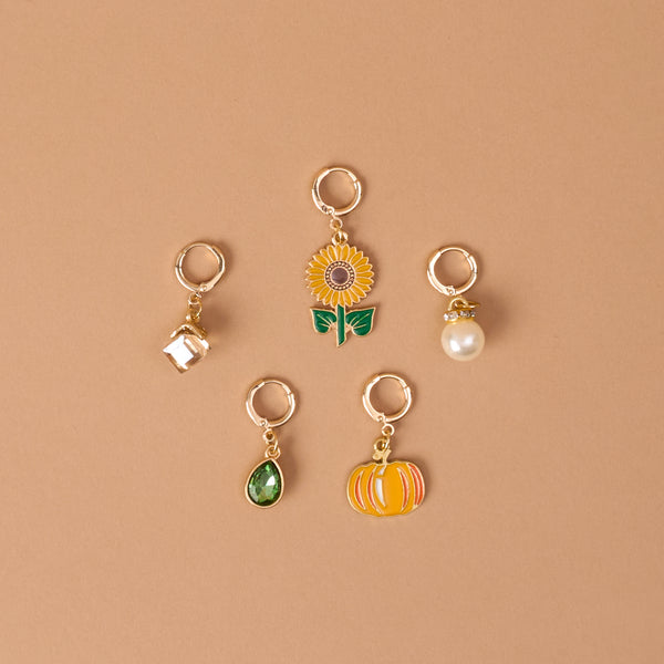 Sunflower Fields The Ultimate Charm Pack by The Darling Effect features five gold keychains—a white house, a sunflower, a pearl, a green teardrop gem, and an orange pumpkin—beautifully arranged on a tan background in a loose circular pattern.