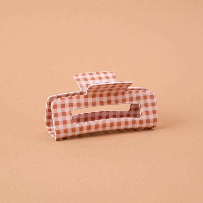 The Maple Gingham Claw Clip by The Darling Effect is a stylish rectangular hair accessory with rounded edges, featuring a charming pink and white gingham checkered pattern and displayed on a plain light brown background.