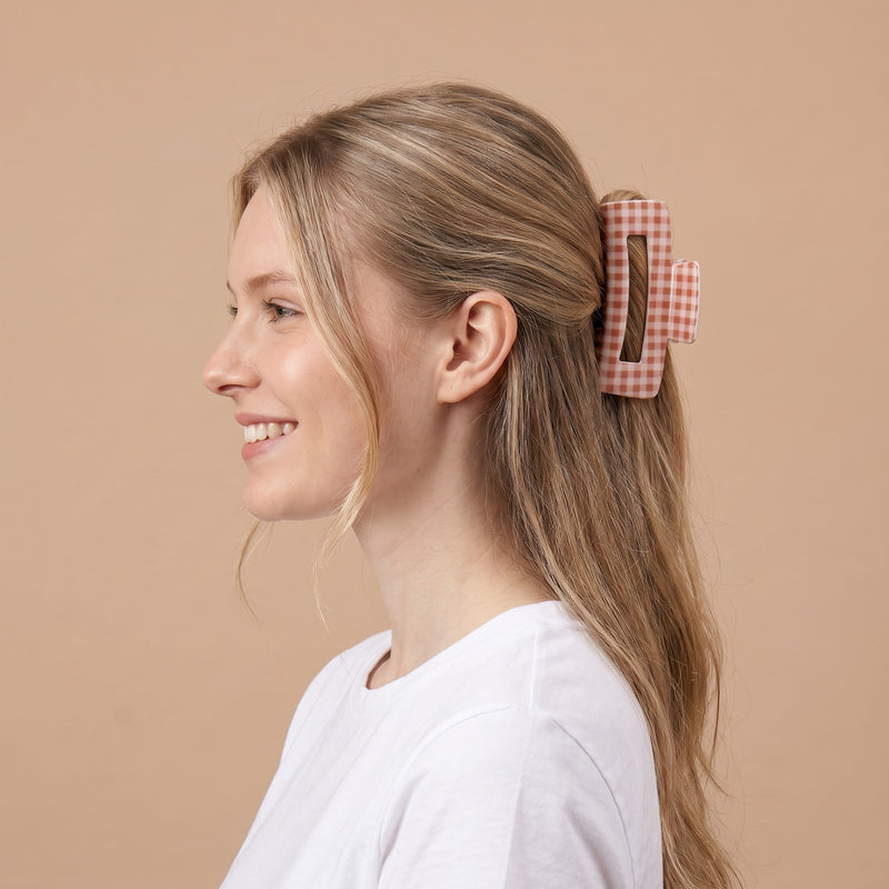 A woman with long, blonde hair, wearing a white shirt, smiles in profile. Her hair is clipped back with The Darling Effect’s stylish Maple Gingham Claw Clip—a large pink and white checkered accessory—set against a beige background.