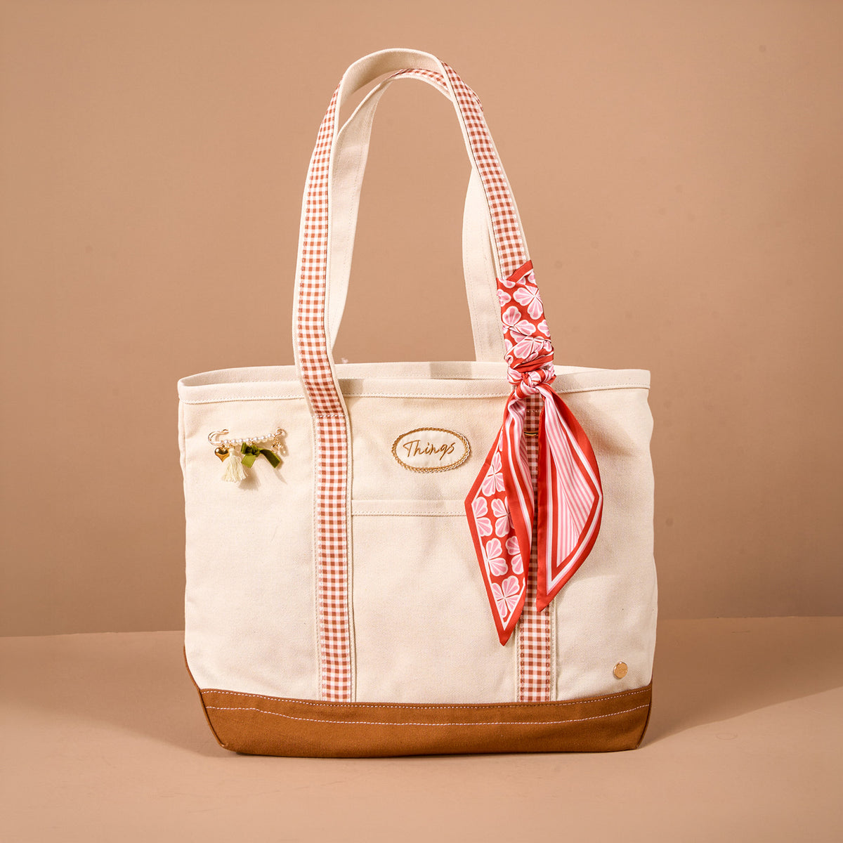 The Maple Gingham The Darling Canvas Tote Bag by The Darling Effect features a cream canvas body with a roomy interior, accented by a brown base, red plaid handles, a red patterned scarf tied to one handle, and a small decorative pin. It is pictured against a plain light brown background.
