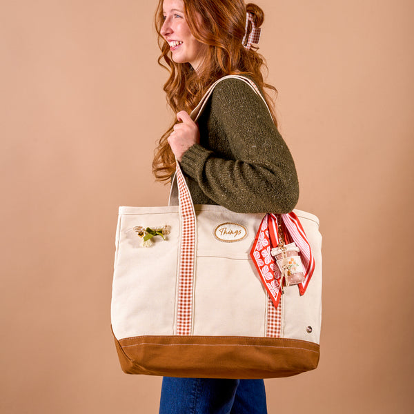 A smiling woman with long red hair holds The Darling Effect’s Maple Gingham The Darling Canvas Tote Bag, adorned with a scarf, floral pin, and an embroidered “Thanks” patch, standing against a beige background.
