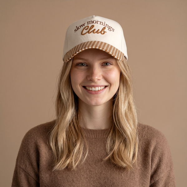 A smiling person wearing The Darling Effect’s Slow Mornings Club Classic Canvas Hat and a brown sweater stands in front of a plain beige background, effortlessly showcasing a stylish everyday look.