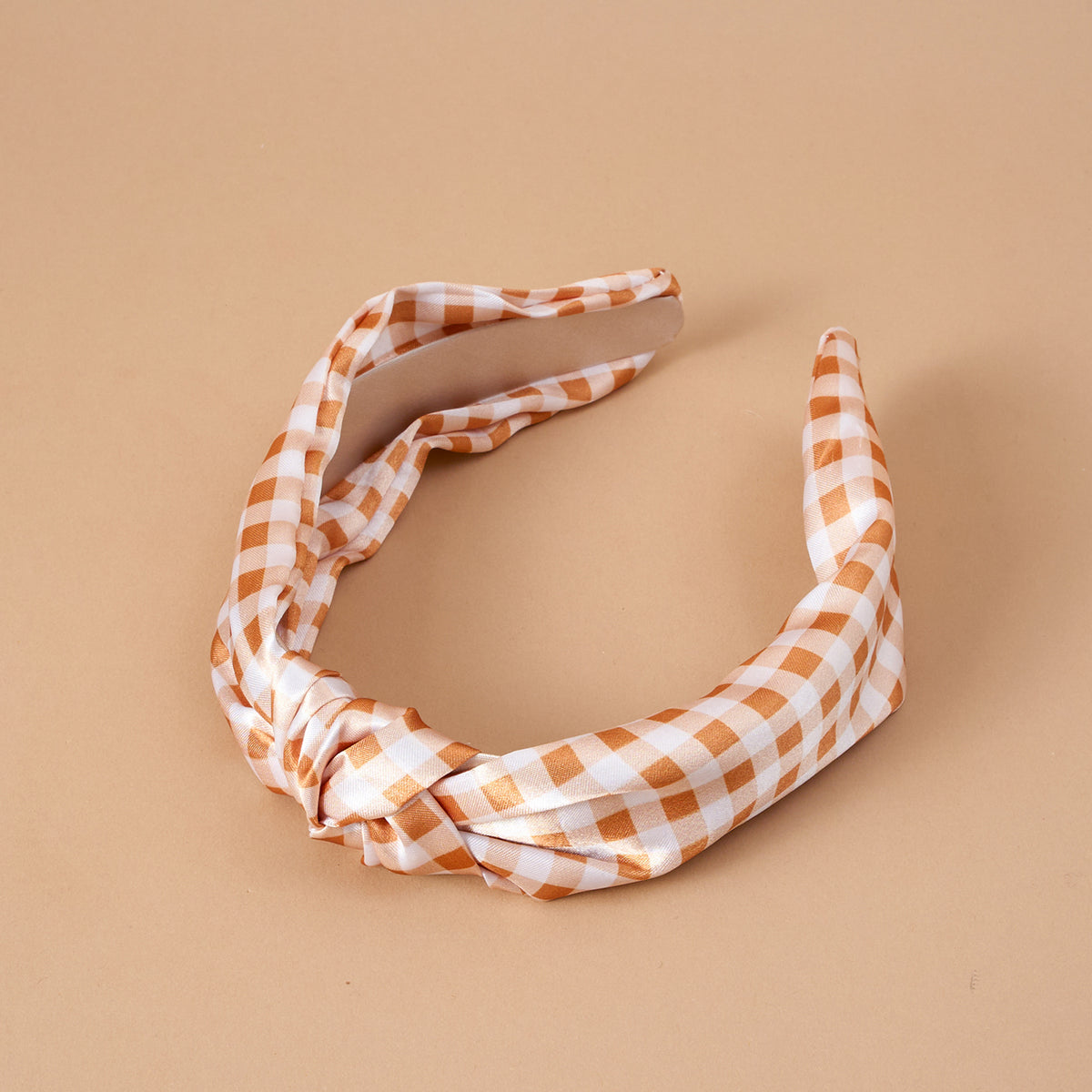 The Maple Gingham Classic Twist Headband by The Darling Effect features a brown and white checkered pattern with a knotted design at the front, displayed on a beige background.