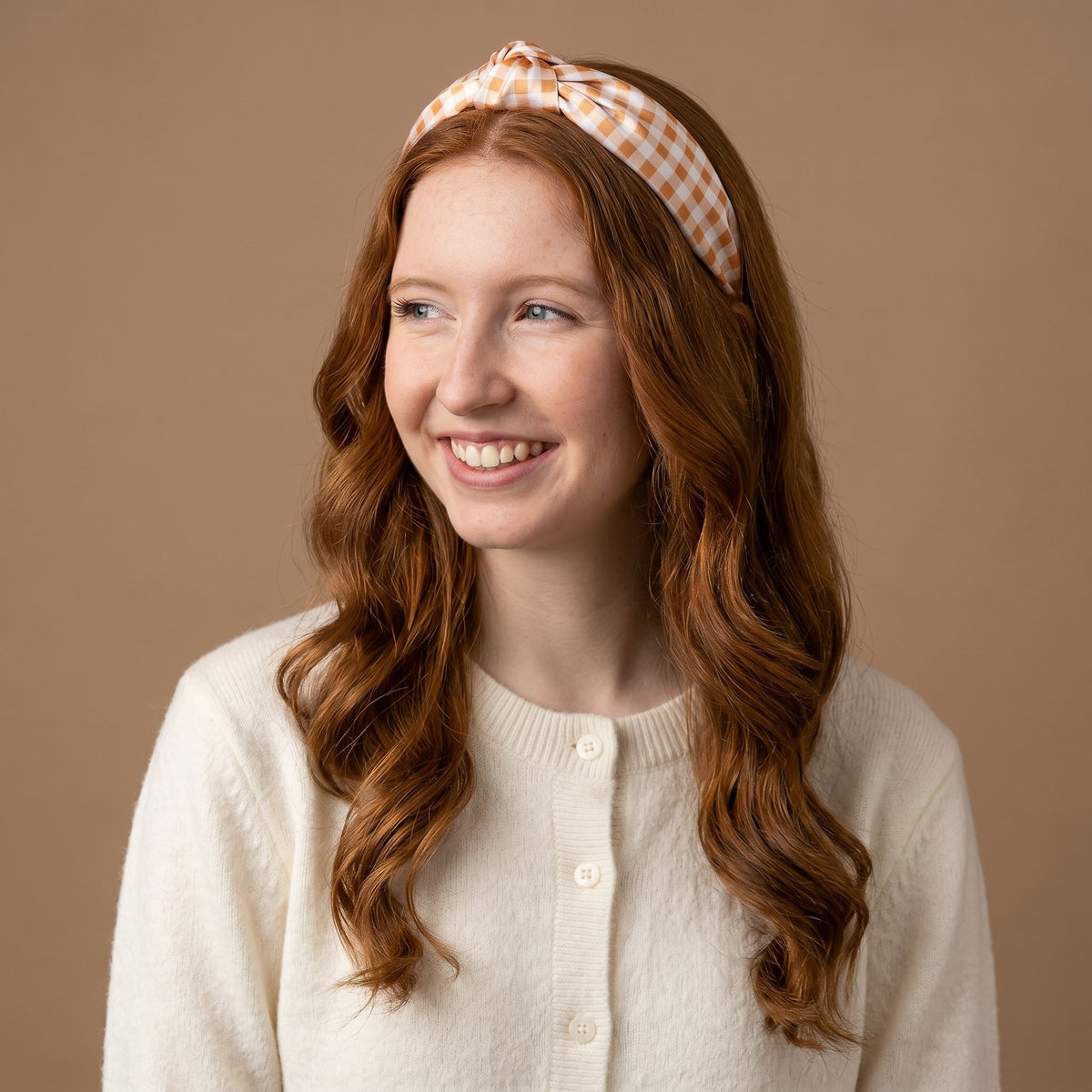 A young woman with long red hair, dressed in a cream sweater and wearing The Darling Effect Maple Gingham Classic Twist Headband, smiles while looking to the side against a plain beige background.
