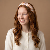 A young woman with long red hair, dressed in a cream sweater and wearing The Darling Effect Maple Gingham Classic Twist Headband, smiles while looking to the side against a plain beige background.