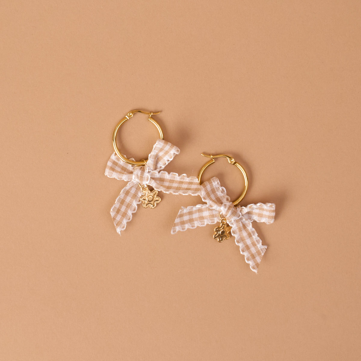 The Darling Effect’s Harvest Honey Signature Hoop Earrings feature two gold hoops embellished with white gingham ribbon bows and small flower-shaped charms, all showcased against a neutral beige background.
