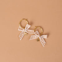 The Darling Effect’s Harvest Honey Signature Hoop Earrings feature two gold hoops embellished with white gingham ribbon bows and small flower-shaped charms, all showcased against a neutral beige background.