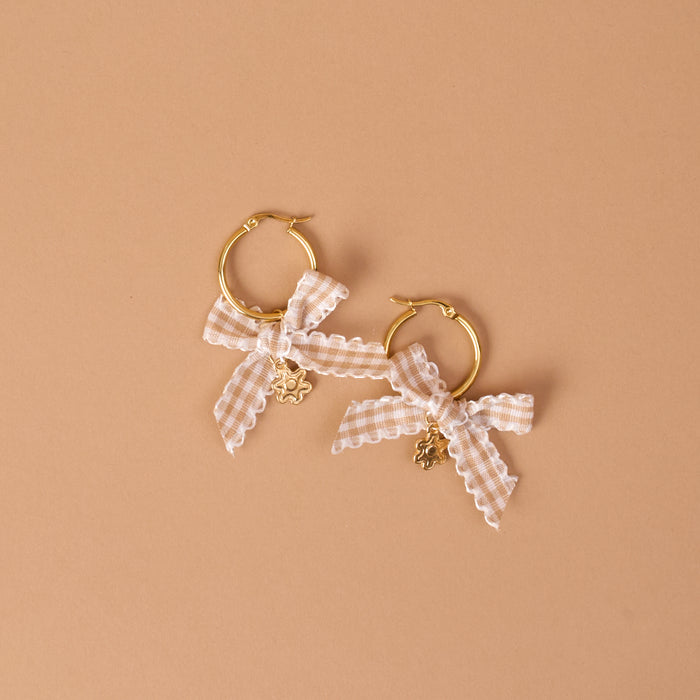 The Darling Effect’s Harvest Honey Signature Hoop Earrings feature two gold hoops embellished with white gingham ribbon bows and small flower-shaped charms, all showcased against a neutral beige background.