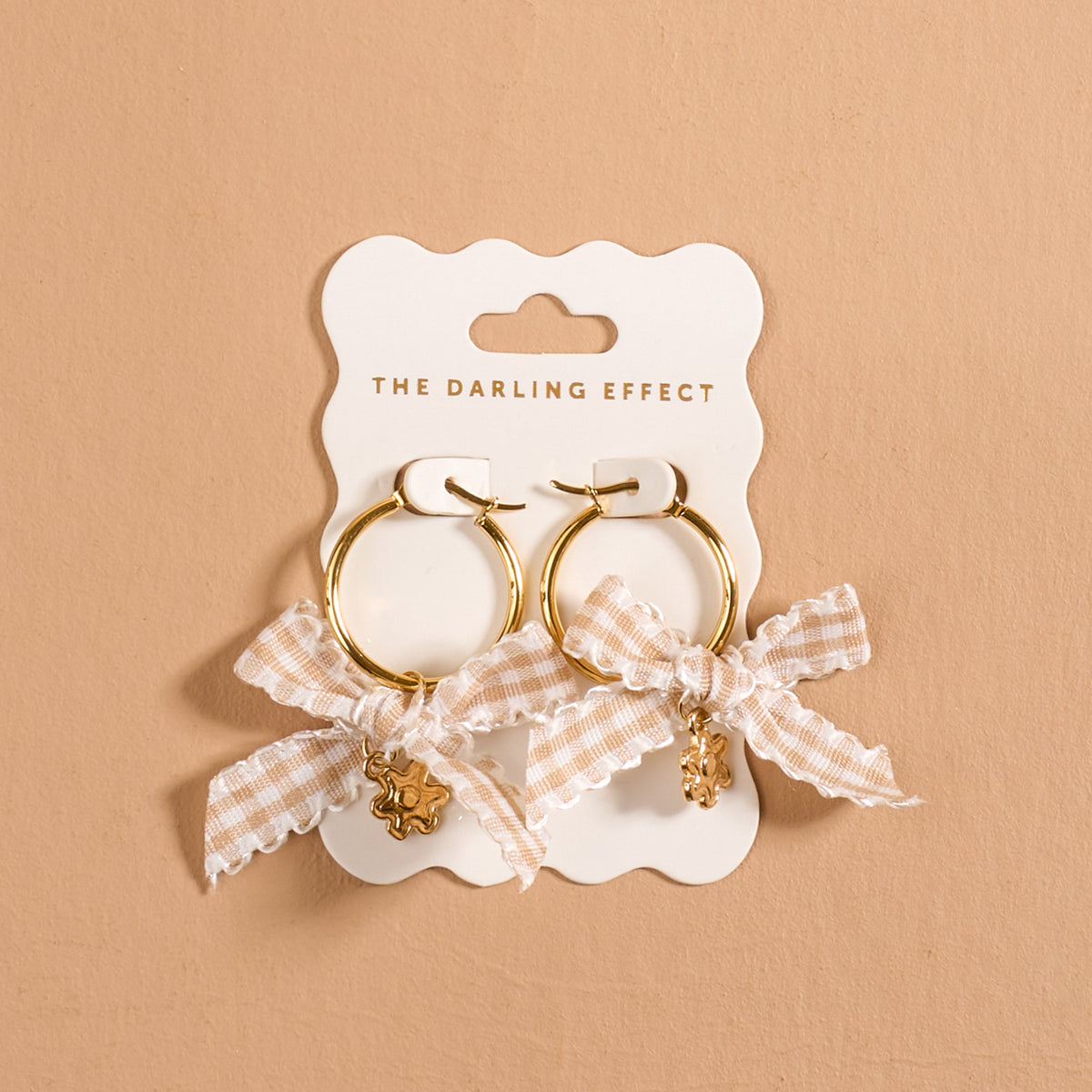 Harvest Honey Signature Hoop Earrings by The Darling Effect feature gold hoops adorned with checkered cream bows and delicate dangling charms, elegantly displayed on a branded card against a neutral beige background. These lightweight earrings beautifully combine playful elegance with modern charm, making them an ideal addition to any collection of charm earrings.
