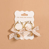 Harvest Honey Signature Hoop Earrings by The Darling Effect feature gold hoops adorned with checkered cream bows and delicate dangling charms, elegantly displayed on a branded card against a neutral beige background. These lightweight earrings beautifully combine playful elegance with modern charm, making them an ideal addition to any collection of charm earrings.