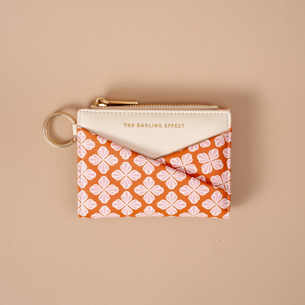 Introducing the Sienna Spice Luxe Pocket Wallet by The Darling Effect—a compact zippered wallet with a key ring, designed with a cream top and an orange base decorated in a white and pink floral pattern. Crafted from vegan leather for both style and sustainability, this pouch features “THE DARLING EFFECT” embossed in gold on the cream section, offering an elegant finishing touch.