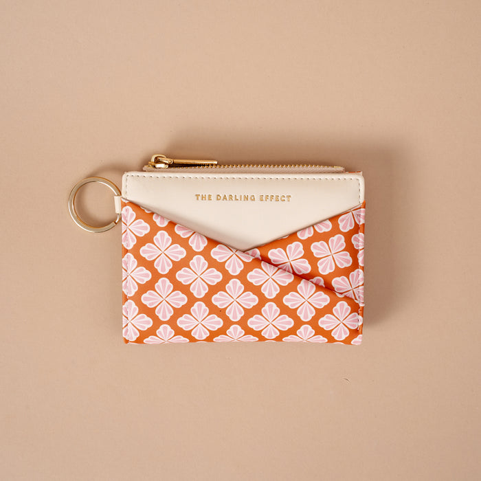 Introducing the Sienna Spice Luxe Pocket Wallet by The Darling Effect—a compact zippered wallet with a key ring, designed with a cream top and an orange base decorated in a white and pink floral pattern. Crafted from vegan leather for both style and sustainability, this pouch features “THE DARLING EFFECT” embossed in gold on the cream section, offering an elegant finishing touch.