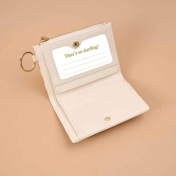 The Darling Effect Sienna Spice Luxe Pocket Wallet, crafted from cream-colored vegan leather and featuring a keyring, is shown open to reveal a card slot with a card that reads “That’s so darling!” against a beige backdrop, highlighted by an elegant criss-cross design.
