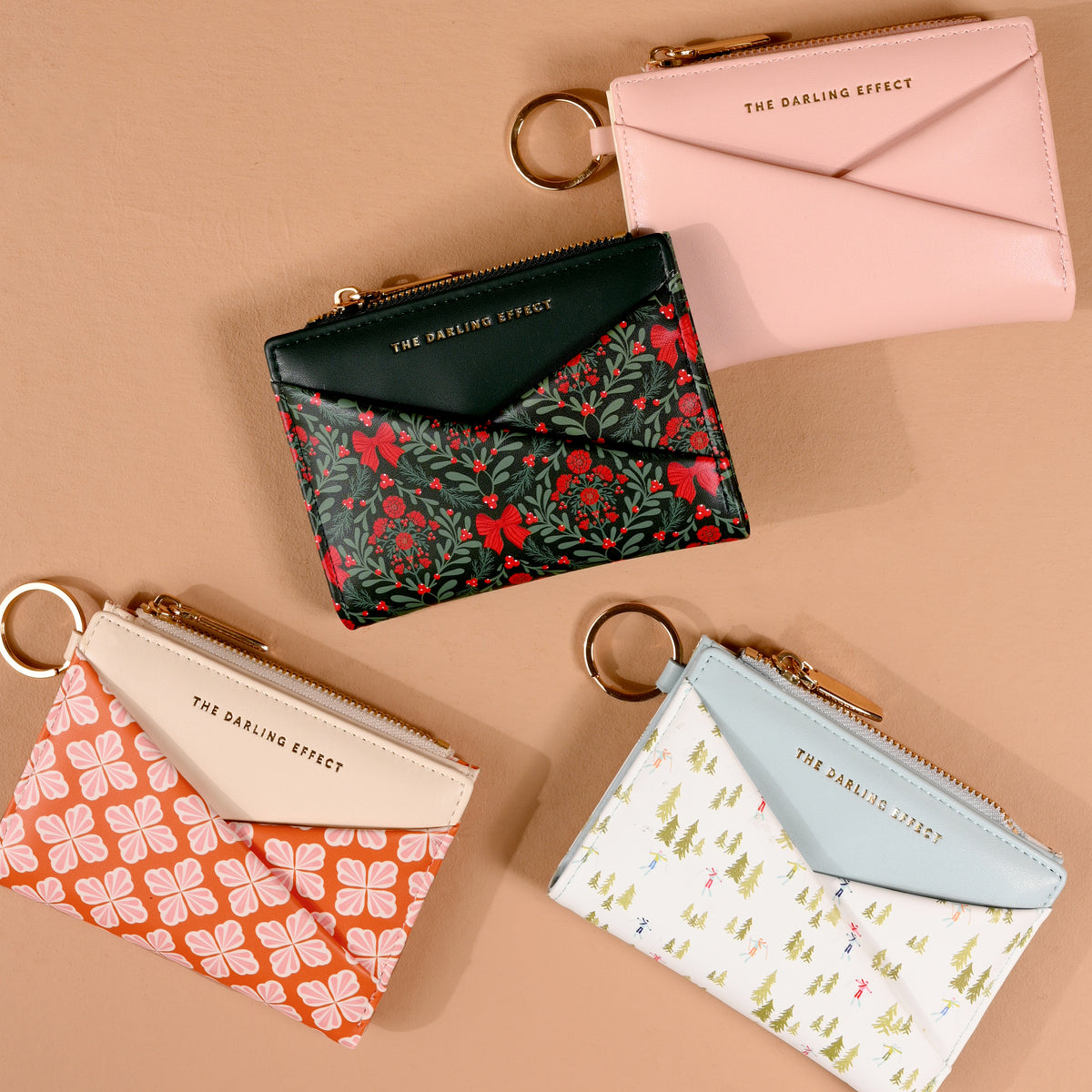 Four Sienna Spice Luxe Pocket Wallets by The Darling Effect are arranged on a tan surface. Each vegan leather wallet features an envelope flap, a keyring, and gold THE DARLING EFFECT text. Designs include floral, geometric, tree, criss-cross patterns, and a plain blush pink option.