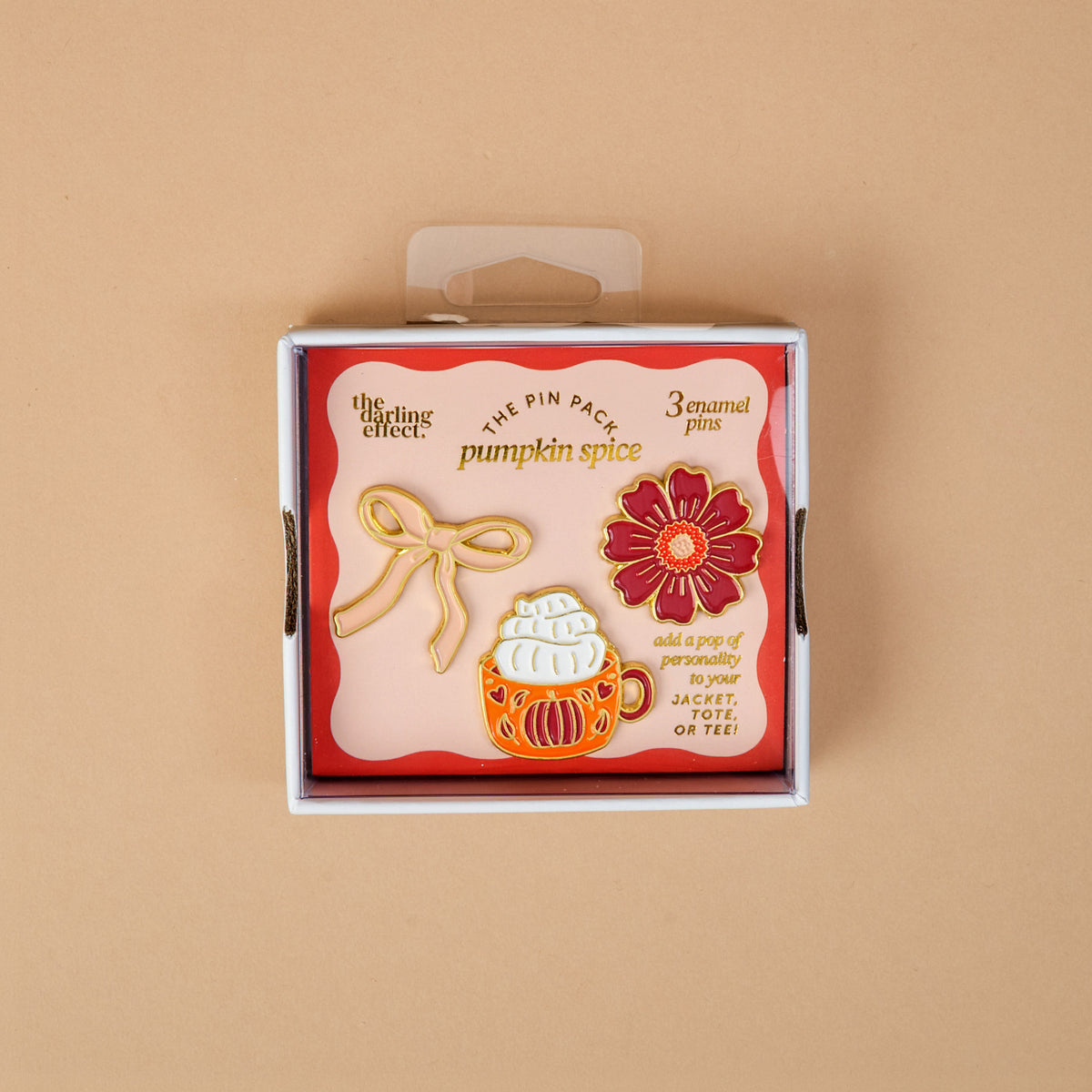 Introducing Pumpkin Spice The Pin Pack by The Darling Effect: a thoughtfully curated set displayed on a peach background, featuring three vibrant pins—a cheerful yellow bow, a cozy pumpkin spice latte, and a bright red flower. The packaging reads “pumpkin spice” and encourages you to add these collectible pins to your favorite jacket or tee.