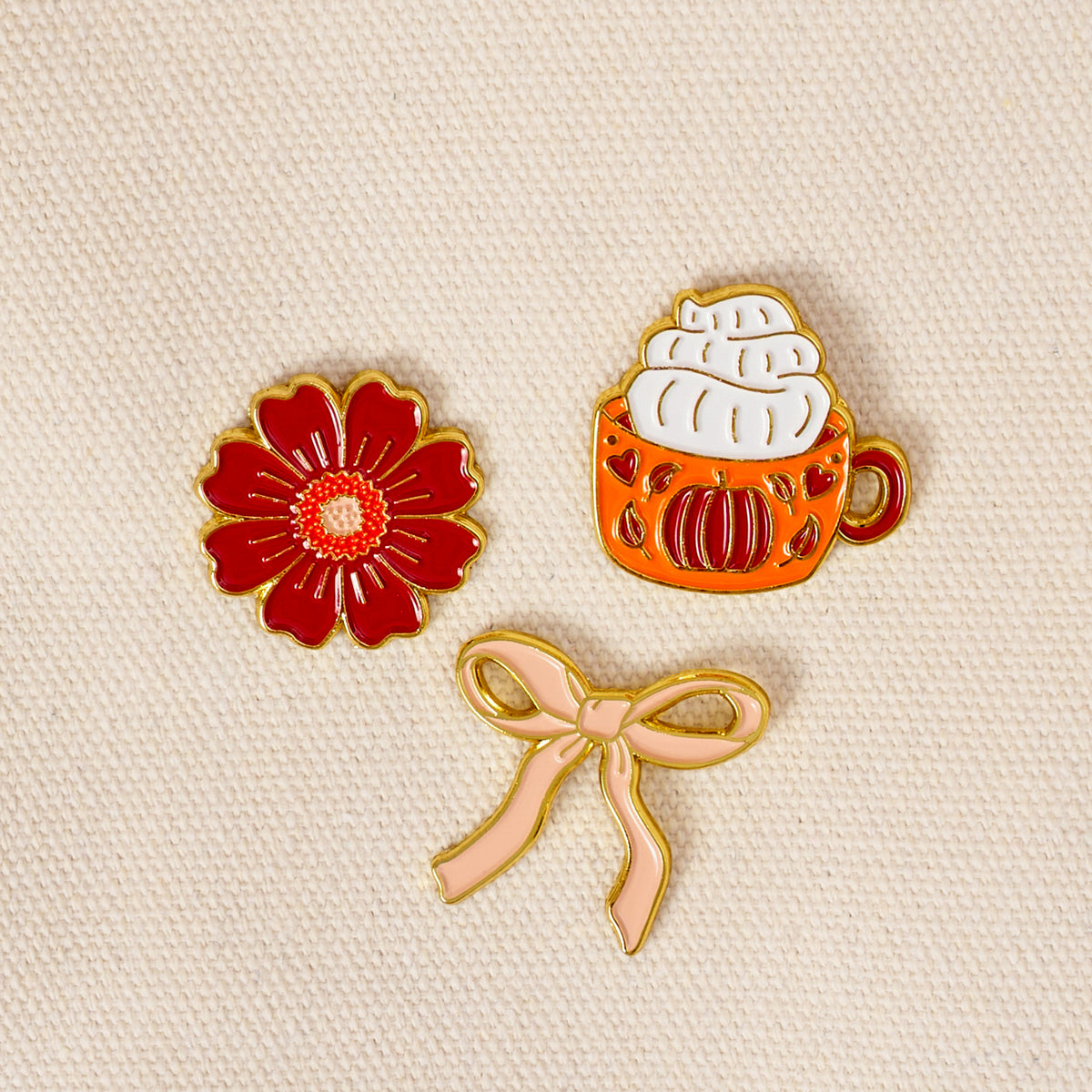 Three expressive pins from The Darling Effect’s Pumpkin Spice The Pin Pack rest on a beige textured fabric: a red flower, an orange mug topped with whipped cream, and a light pink bow. This curated set is perfect for anyone looking to add charm and personality to their pin collection.