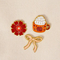 Three expressive pins from The Darling Effect’s Pumpkin Spice The Pin Pack rest on a beige textured fabric: a red flower, an orange mug topped with whipped cream, and a light pink bow. This curated set is perfect for anyone looking to add charm and personality to their pin collection.