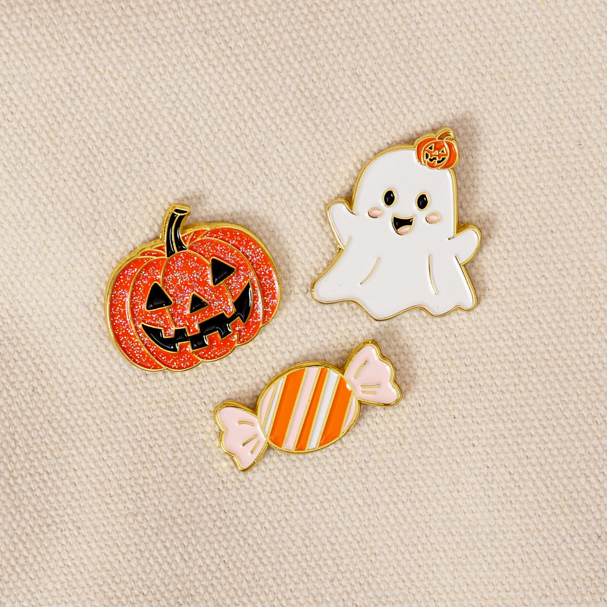 Showcased on beige fabric, the Trick or Treat The Pin Pack by The Darling Effect features three charming enamel pins: a glittery orange jack-o-lantern, a smiling white ghost topped with a tiny pumpkin, and an orange-and-white striped candy. This playful set is ideal for anyone who loves expressive accessories or collecting pins.