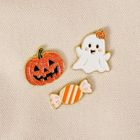Showcased on beige fabric, the Trick or Treat The Pin Pack by The Darling Effect features three charming enamel pins: a glittery orange jack-o-lantern, a smiling white ghost topped with a tiny pumpkin, and an orange-and-white striped candy. This playful set is ideal for anyone who loves expressive accessories or collecting pins.