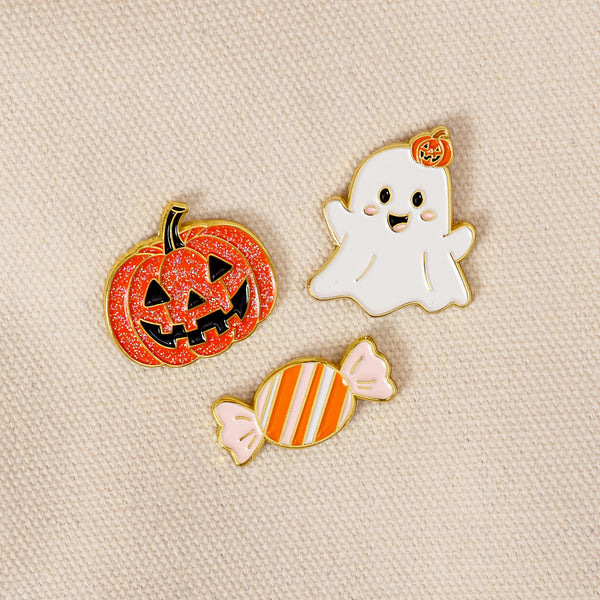 Showcased on beige fabric, the Trick or Treat The Pin Pack by The Darling Effect features three charming enamel pins: a glittery orange jack-o-lantern, a smiling white ghost topped with a tiny pumpkin, and an orange-and-white striped candy. This playful set is ideal for anyone who loves expressive accessories or collecting pins.