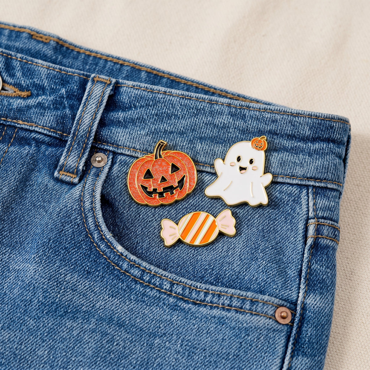 Three collectible pins from The Darling Effect’s Trick or Treat The Pin Pack—a jack-o-lantern, a smiling ghost, and a striped candy—are attached to the small pocket of blue denim jeans. These expressive accessories stand out against the light-colored fabric background.