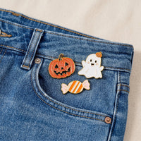 Three collectible pins from The Darling Effect’s Trick or Treat The Pin Pack—a jack-o-lantern, a smiling ghost, and a striped candy—are attached to the small pocket of blue denim jeans. These expressive accessories stand out against the light-colored fabric background.