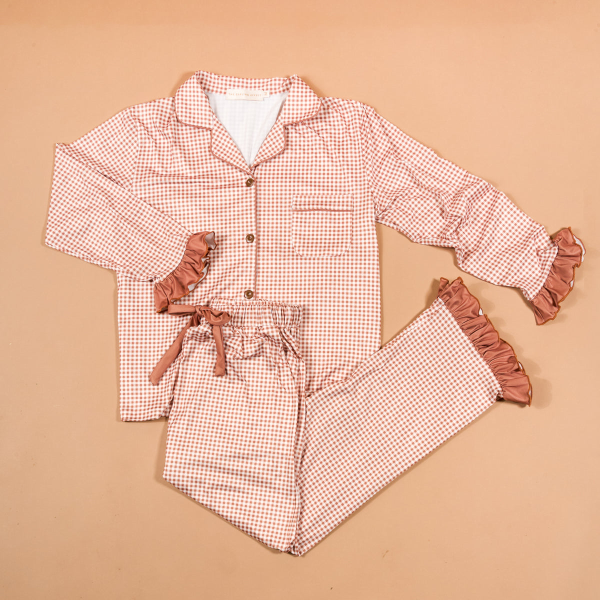 Experience cozy comfort and charming style with the Maple Gingham Sweet Dreams Pajama Pants Set from The Darling Effect. This pink and white checkered set features a long-sleeve button-up top paired with matching pants, both highlighted by playful ruffled cuffs for a feminine finish. The pants include a comfortable drawstring waist, making this set perfect for relaxing nights in or as an adorable gift option. Displayed on a beige background.