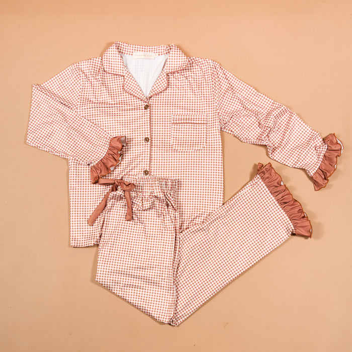 Experience cozy comfort and charming style with the Maple Gingham Sweet Dreams Pajama Pants Set from The Darling Effect. This pink and white checkered set features a long-sleeve button-up top paired with matching pants, both highlighted by playful ruffled cuffs for a feminine finish. The pants include a comfortable drawstring waist, making this set perfect for relaxing nights in or as an adorable gift option. Displayed on a beige background.