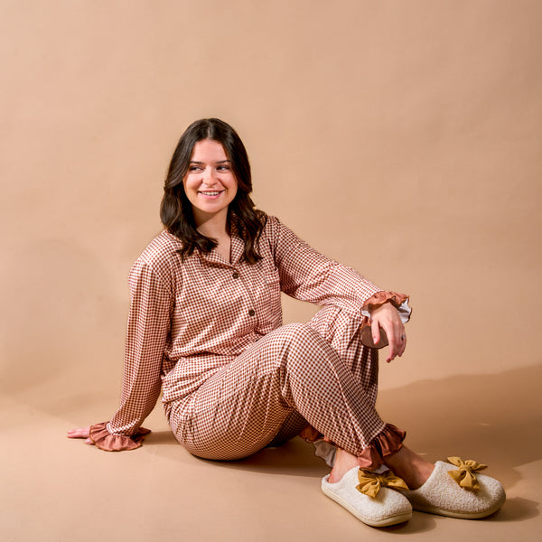 A woman in the Maple Gingham Sweet Dreams Pajama Pants Set from The Darling Effect sits on the floor against a beige background, smiling. She has dark hair and is wearing cozy bow slippers, perfect for relaxing in your favorite comfy loungewear.