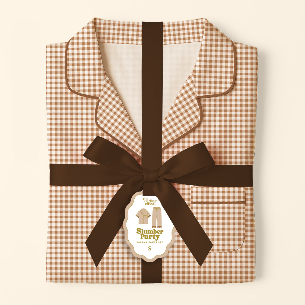 Neatly folded brown and white gingham pajamas with black piping, tied with a dark brown ribbon and a tag reading “Maple Gingham Sweet Dreams Pajama Pants Set” from The Darling Effect—these cozy loungewear pieces make the perfect gift pajama sets for any occasion.