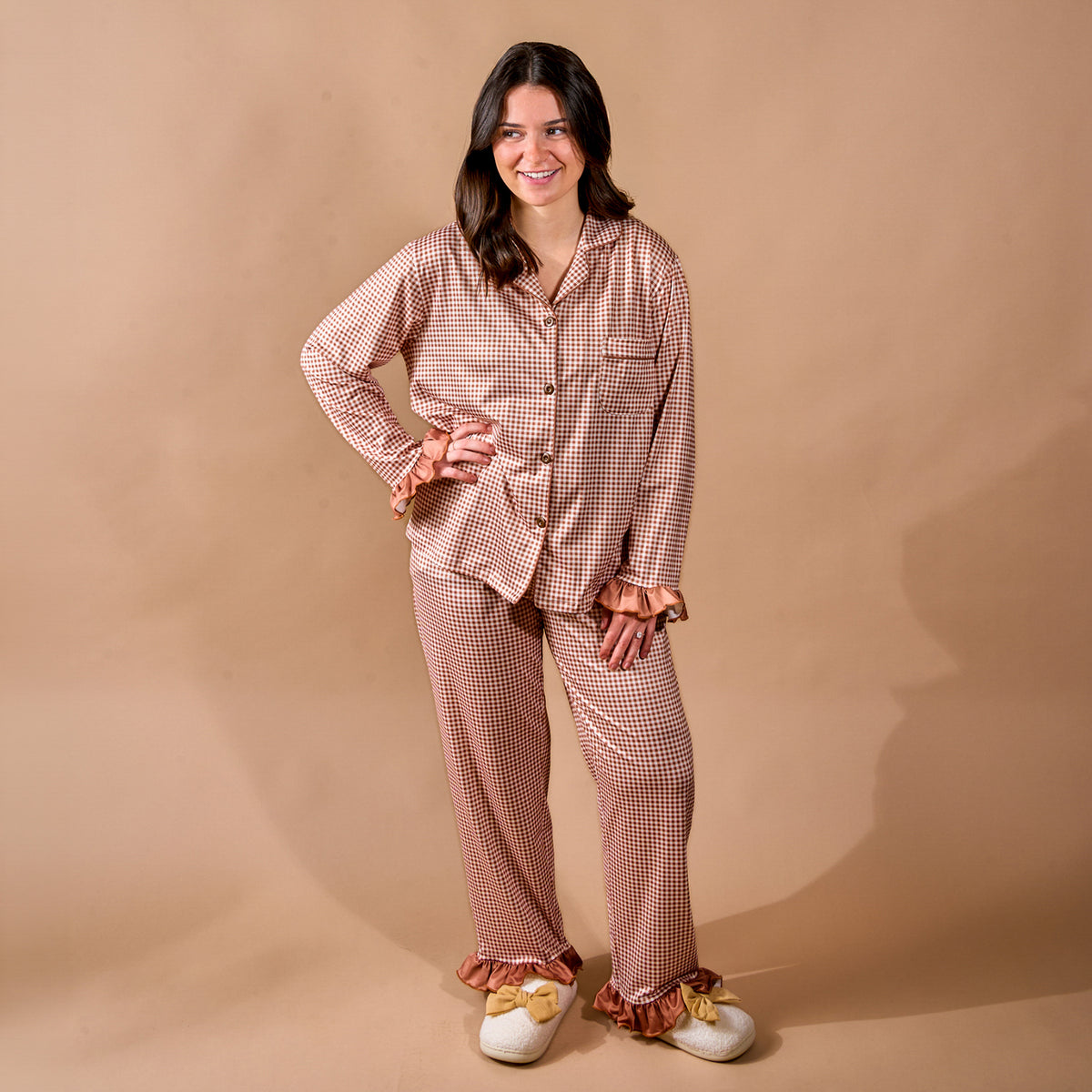 A woman stands smiling, wearing The Darling Effect’s Maple Gingham Sweet Dreams Pajama Pants Set—featuring pink and brown checks with ruffle details—and white bow slippers against a beige background, the perfect cozy loungewear for relaxing at home or gifting to loved ones.