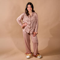 A woman stands smiling, wearing The Darling Effect’s Maple Gingham Sweet Dreams Pajama Pants Set—featuring pink and brown checks with ruffle details—and white bow slippers against a beige background, the perfect cozy loungewear for relaxing at home or gifting to loved ones.
