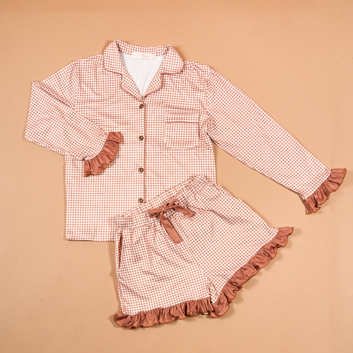 Displayed on a beige background, the Maple Gingham Sweet Dreams Pajama Shorts Set by The Darling Effect features a pink and white checkered long-sleeve top with buttons and matching shorts, both accented with pink ruffles—offering the perfect blend of style and comfort for your nightly routine.