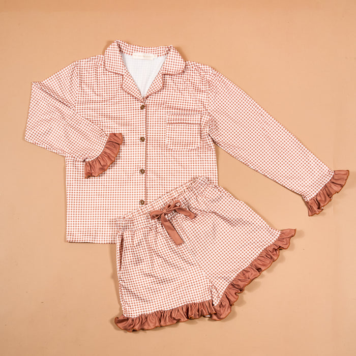 Displayed on a beige background, the Maple Gingham Sweet Dreams Pajama Shorts Set by The Darling Effect features a pink and white checkered long-sleeve top with buttons and matching shorts, both accented with pink ruffles—offering the perfect blend of style and comfort for your nightly routine.