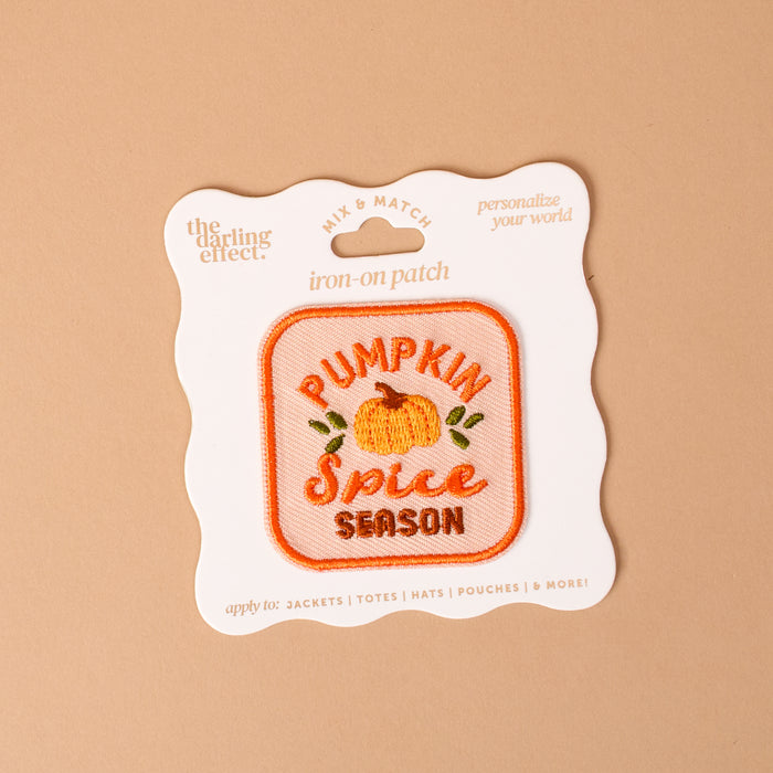 The Darling Effect’s On Patch-Pumpkin Spice Season Mix & Match Iron is a square iron-on patch featuring an orange pumpkin and the phrase “Pumpkin Spice Season” embroidered on it, displayed on beige packaging. Perfect for customizing clothing or accessories, this patch adds a cozy touch to any colorful collection.