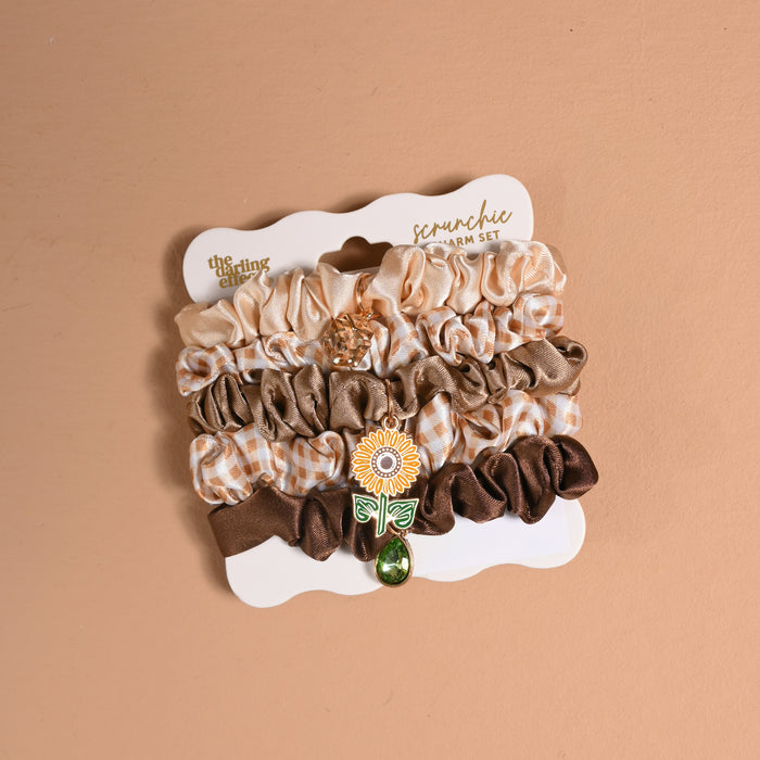 The Maple Gingham Scrunchie Charm Set by The Darling Effect includes five stylish brown and beige scrunchies, featuring a sunflower hair charm and a bead. Displayed on a white card against a light brown background, this set is perfect for adding trendy flair to your collection of hair accessories.