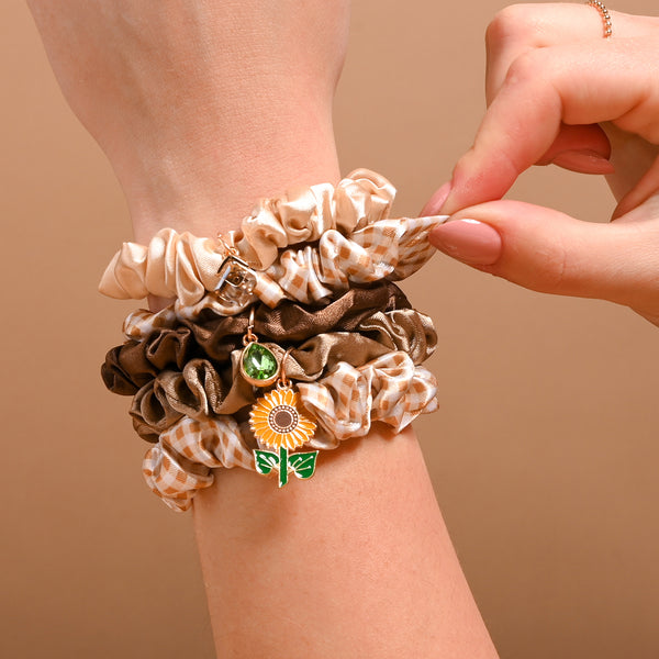 A hand adjusts a stack of stylish hair accessories from The Darling Effect’s Maple Gingham Scrunchie Charm Set, featuring beige, brown, and plaid scrunchies—one adorned with a sunflower and leaf charm—elegantly displayed against a plain brown background.