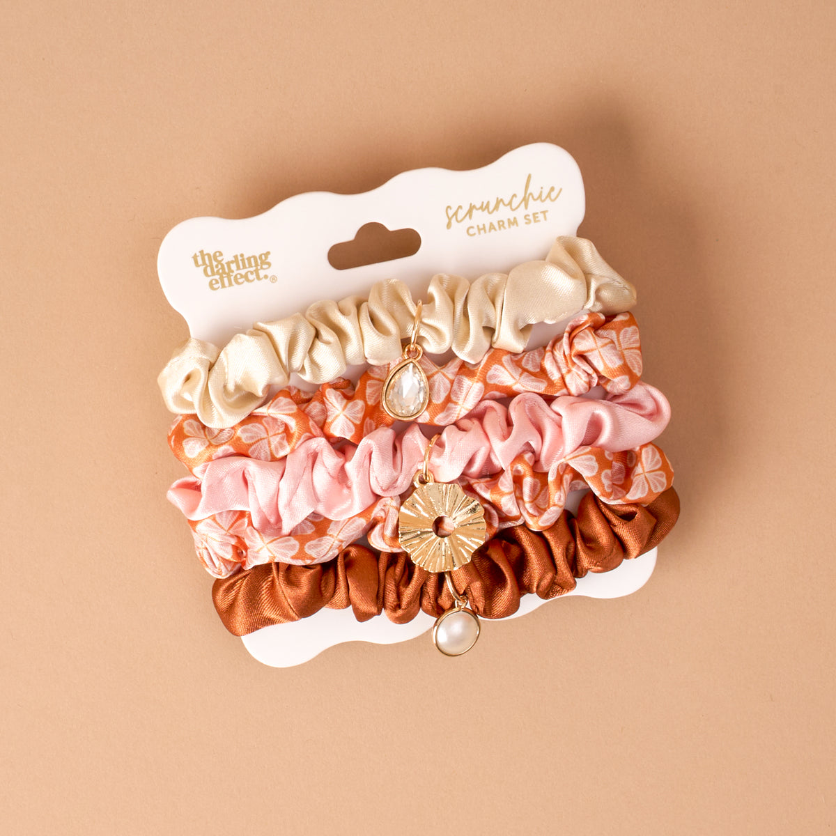The Sienna Spice Scrunchie Charm Set by The Darling Effect features three decorative scrunchies in cream, pink floral, and copper, each embellished with gold hair charms. Presented on white packaging labeled “scrunchie charm set” against a beige background, it’s the ideal choice for anyone looking for stylish hair accessories.