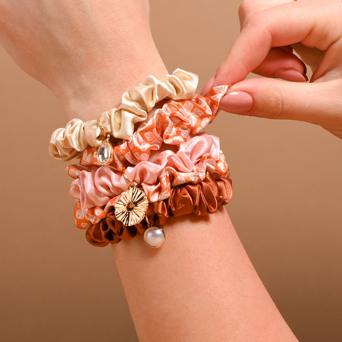 A wrist stacked with The Darling Effect’s Sienna Spice Scrunchie Charm Set—mini satin scrunchies in beige, pink, rust, and floral orange, some embellished with gold and pearl hair charms. A hand adjusts these stylish accessories against a warm beige background.