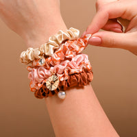 A wrist stacked with The Darling Effect’s Sienna Spice Scrunchie Charm Set—mini satin scrunchies in beige, pink, rust, and floral orange, some embellished with gold and pearl hair charms. A hand adjusts these stylish accessories against a warm beige background.