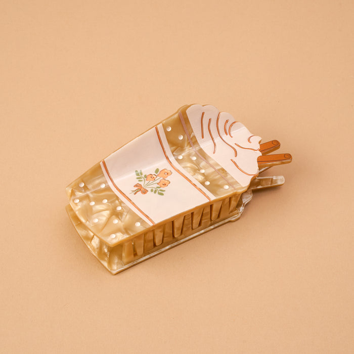 Introducing the “Coffee Run Shape Up Claw Clip” by The Darling Effect—a rectangular, cream-colored hair accessory adorned with a whimsical cake slice design. Featuring delightful white icing, playful polka dots, floral accents, and two charming brown sticks on top, this claw clip is perfect for adding a touch of sweetness to any cute hairstyle. Presented against a plain beige background.
