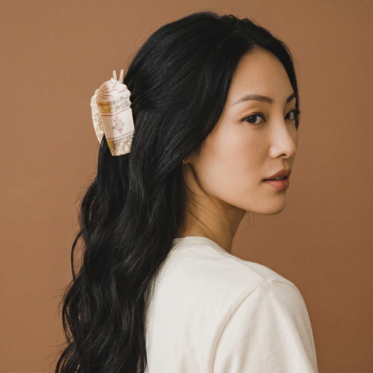 A woman with long black hair and a white shirt faces sideways, wearing The Darling Effect’s Coffee Run Shape Up Claw Clip—a charming hair accessory shaped like a porcelain teacup—against a brown background.