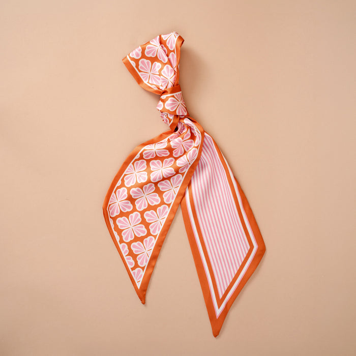 The Sienna Spice Twirl & Tie Scarf Accessory by The Darling Effect showcases vibrant orange and white floral and striped patterns, tied in a loose knot and displayed on a plain beige background, offering endless styling possibilities.
