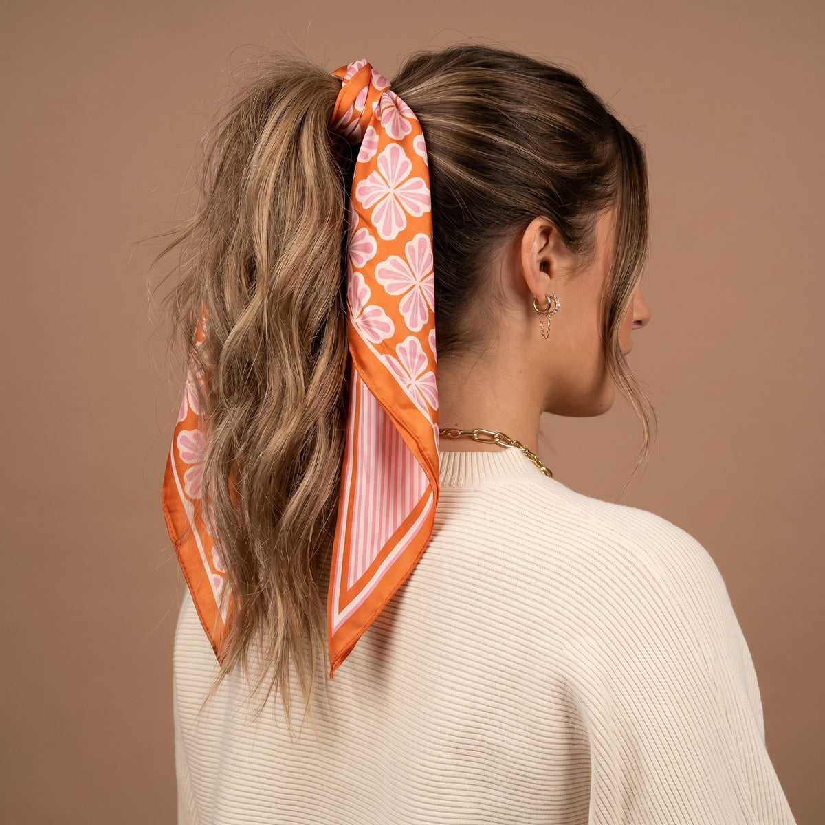 A woman with wavy brown hair styled in a ponytail, secured with The Darling Effect Sienna Spice Twirl & Tie Scarf Accessory, wears a cream sweater and gold jewelry as she faces away from the camera against a beige background.